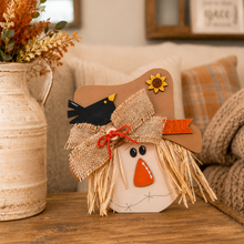 Load image into Gallery viewer, A fall-themed decorative scarecrow sign on a wooden table next to a rustic ceramic vase with dried flowers. The scarecrow has a tan hat with a yellow sunflower, a black bird, a burlap bow, and a white face with a stitched orange triangular nose.