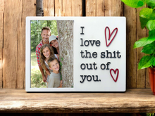 Load image into Gallery viewer, This image features a bold and humorous picture frame, perfect for displaying a special memory. The frame, made of pine wood, has a clean white background and is adorned with the playful text "I Love The Shit Out of You" with heart symbols.
