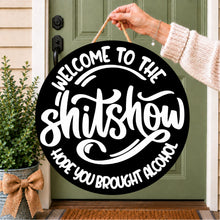Load image into Gallery viewer, A round black hanging sign with white text reading “WELCOME TO THE shitshow HOPE YOU BROUGHT ALCOHOL,” held by a person’s hand in front of a green front door. The sign has a rope handle and is shown outdoors on a porch with a potted plant to the left.
