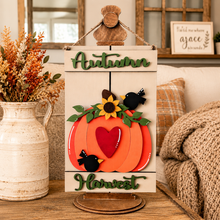 Load image into Gallery viewer, Fall-themed wooden hanging sign with 'Autumn' at the top and 'Harvest' at the bottom, featuring layered orange pumpkins, a red heart, green leaves, yellow sunflower, and black birds on a styled indoor table setting.
