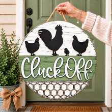 Load image into Gallery viewer, A round wooden hanging sign with a rustic farmhouse design, held by a person's hand in front of a green front door. The sign features black silhouette cutouts of chickens, white script text reading 'Cluck Off', and a distressed white and green wood background.
