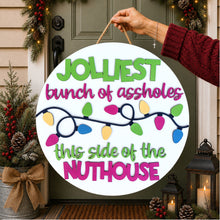 Load image into Gallery viewer, A round white holiday sign with colorful Christmas lights and the text 'JOLLIEST bunch of assholes this side of the NUTHOUSE' being held by a hand in front of a decorated front door.