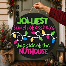 Load image into Gallery viewer, A round black holiday sign held by a hand in front of a decorated front door. The sign reads 'JOLLIEST bunch of assholes this side of the NUTHOUSE' in green and pink lettering with a string of multicolored Christmas lights graphic.