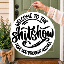 Load image into Gallery viewer, A round white hanging sign with black lettering that reads “WELCOME TO THE shitshow HOPE YOU BROUGHT ALCOHOL,” held by a person’s hand in front of a green front door.

