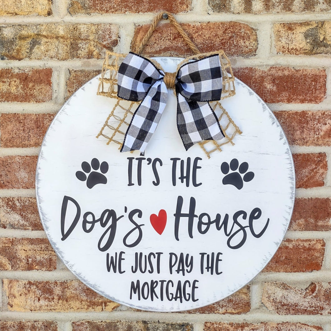 Painted round wood sign that says It's the dog's house, we just pay the mortgage.